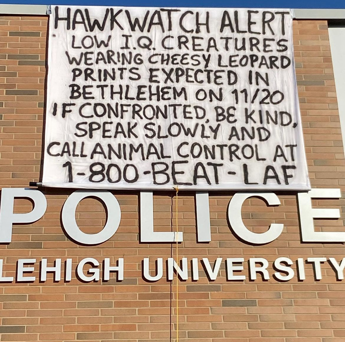 A sign reading "Hawkwatch alert: Low I.Q. creatures wearing cheesy leopard prints expected in Bethlehem on 11/20. If confronted, be kind, speak slowly and call animal control at 1-800-BEAT-LAF" hangs on the side of the Lehigh University Police Department station. 