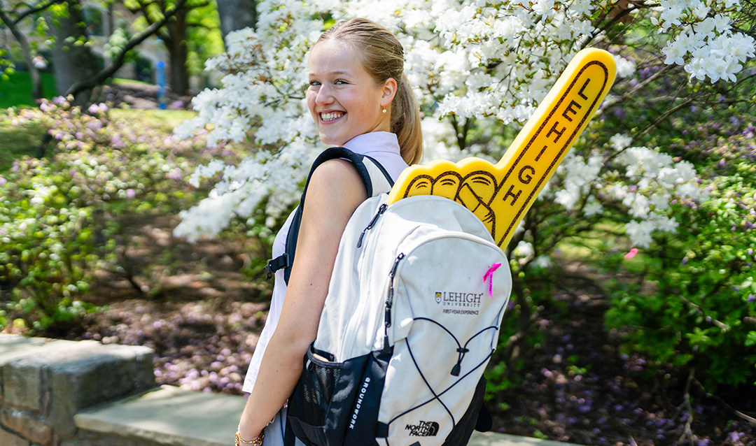 A smiling girl wearing a white shirt and blue overalls stands outdoors with a large white backpack that has a yellow slide attached, in a park-like setting with greenery and flowers in the background.