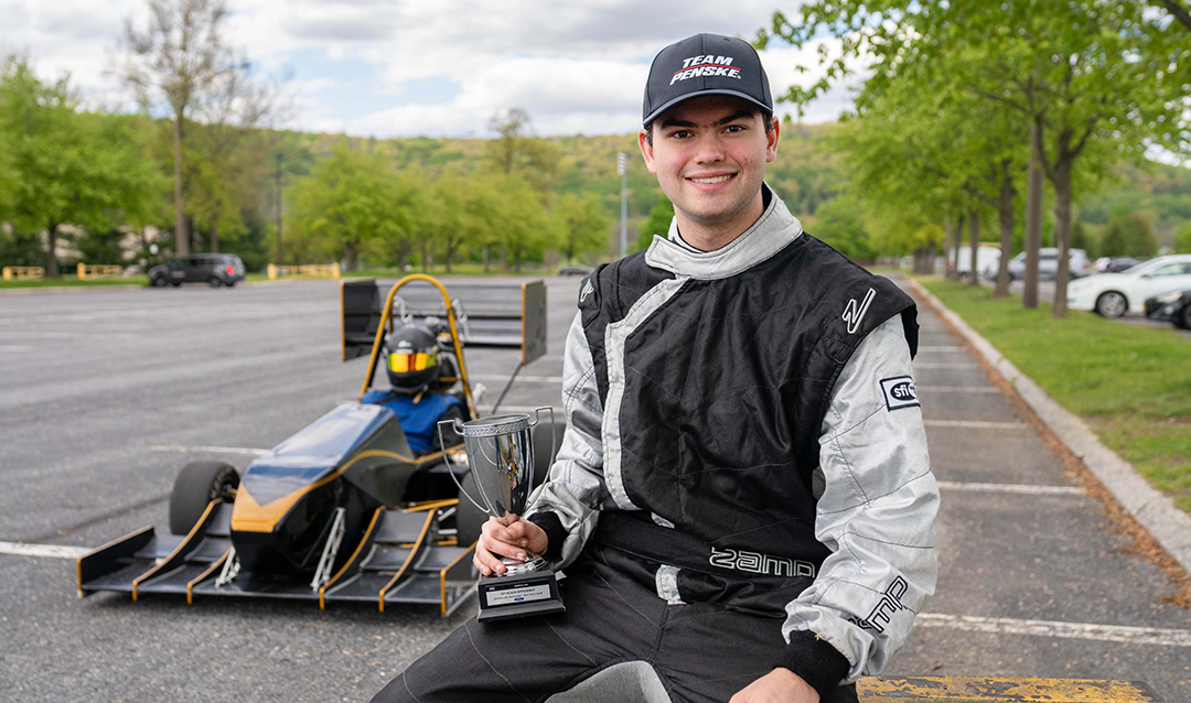 A person in a racing suit and cap sits on a curb, smiling and giving a thumbs up, with a small open-wheel race car parked on the road behind them. Trees and parked cars are visible in the background.