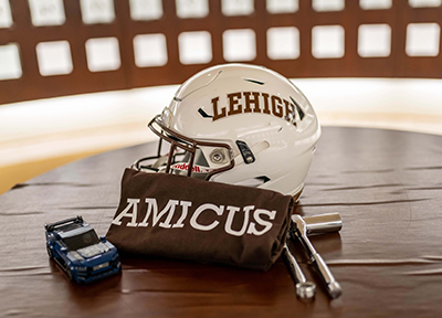 A white Lehigh football helmet, a brown shirt that says AMICUS and a Lego car sitting on a table